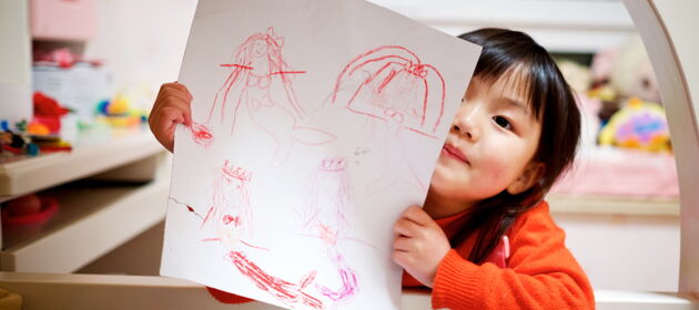 Ein kleines Mädchen in der Tagesbetreuung hält ein Blatt Papier auf dem es gemalt hat hoch