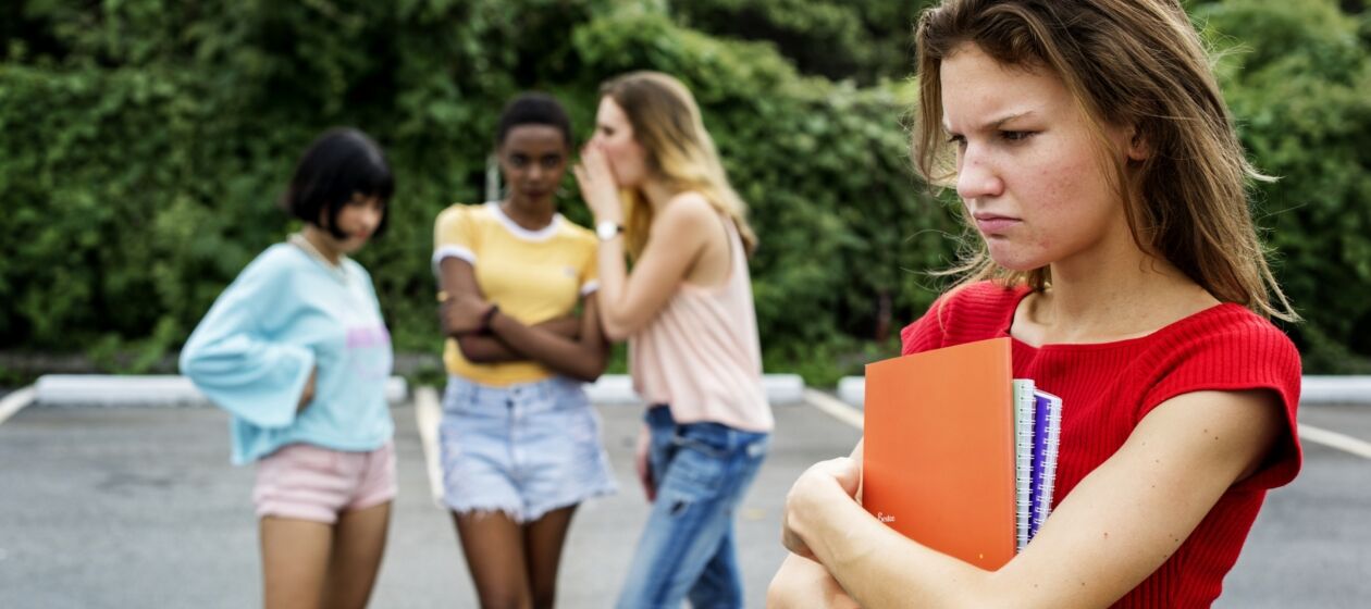 Eine Schülerin mit verärgertem Gesichtsausdruck steht alleine auf dem Schulhof, im Hintergrund reden andere Schülerinnen über sie