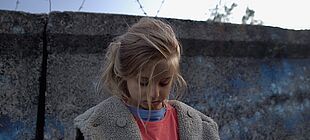 Ein Mädchen steht traurig vor einer Mauer auf der Stacheldraht zu sehen ist