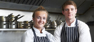 Mann und junge Frau stehen in Küche vor Kochtöpfen