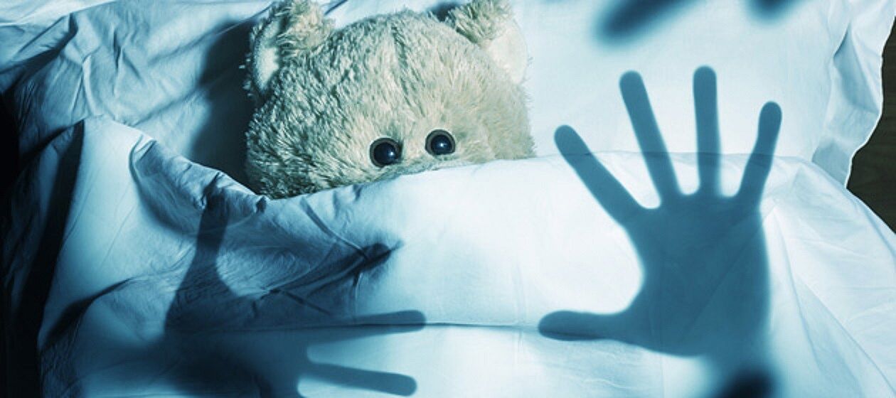 Teddy guckt aus Bettdecke hevor, über ihm der Schatten zweier Hände