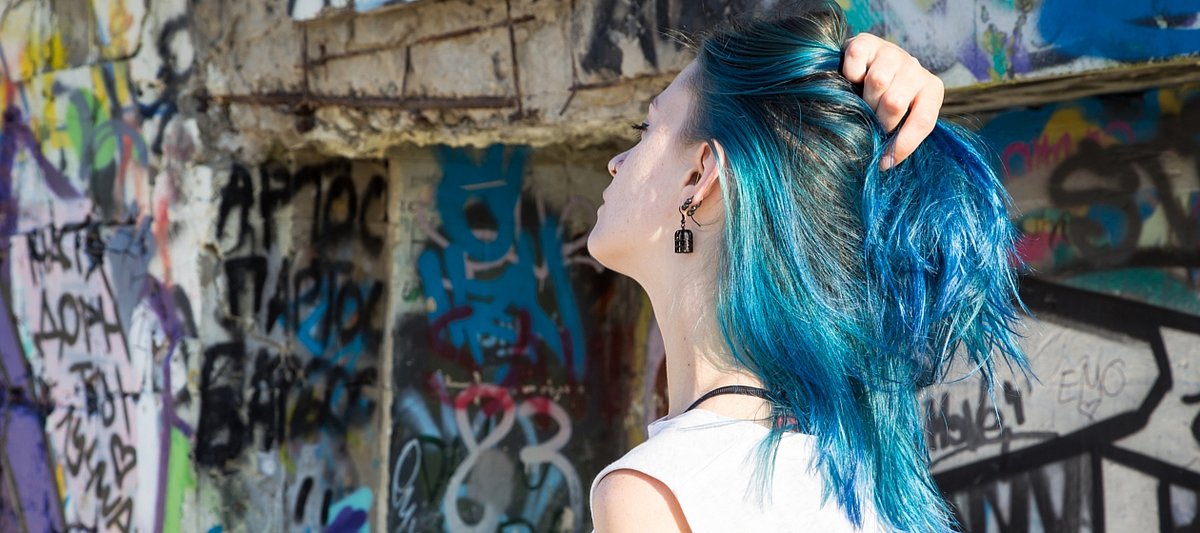 Ein Mädchen fährt sich mit der Hand durch ihre blauen Haaren udn schaut auf eine Mauer mit Graffiti