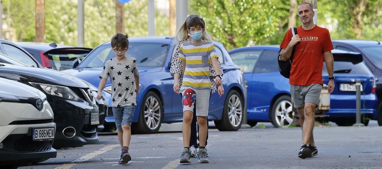 Eine Familie spaziert in einer südeuropäischen Stadt an parkenden Autos vorbei. Es ist Sommer, sie tragen Atemschutzmasken.