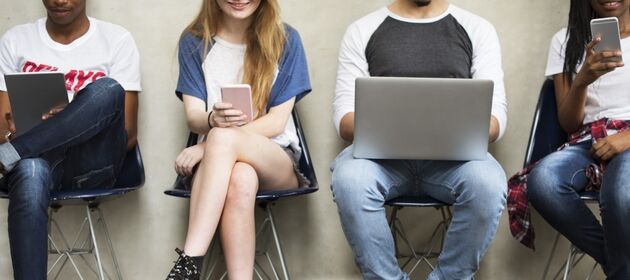 Vier Jugendliche sitzen in einer Stuhlreihe und haben Laptops und Smartphones in den Händen