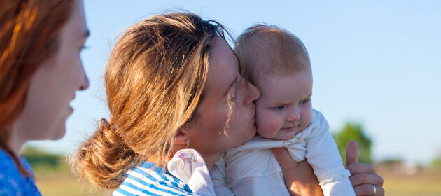 Zwei lesbische Frauen, wobei die eine ein Baby auf dem Arm trägt und küsst. 