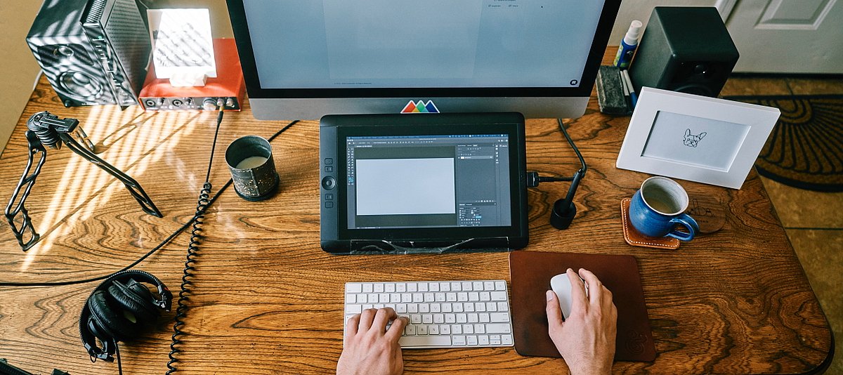 Eine Person sitzt an einem Schreibtisch, der mit unterschiedlichen technischen Geräten bestückt ist, wie einer Tastatur, einem Bildschirm und einem Kopfhörer