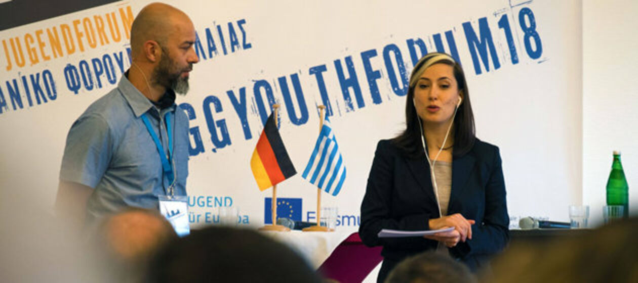 Die beiden Moderator/-innen des Jugendforums an Stehtischen mit einer deutschen und griechischen Flagge. 