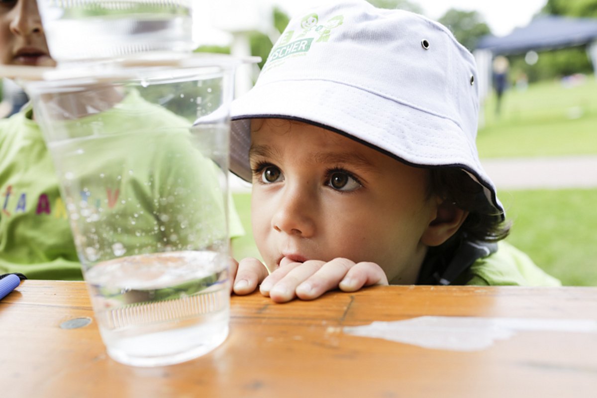 Ein Kind schaut neugierig zu einem Wasserglas hoch, das auf einem Tisch steht