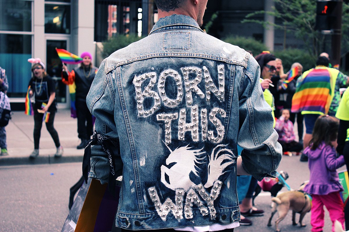 Ein junger Mann trägt eine Jeansjacke mit der Aufschrift "Born this", im Hintergrund stehen Menschen mit Regenbogenflaggen.