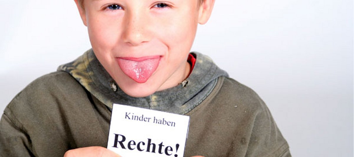 Ein kleiner Junge hält ein Schild mit dem Schriftzug "Kinder haben Rechte" hoch und streckt die Zunge raus