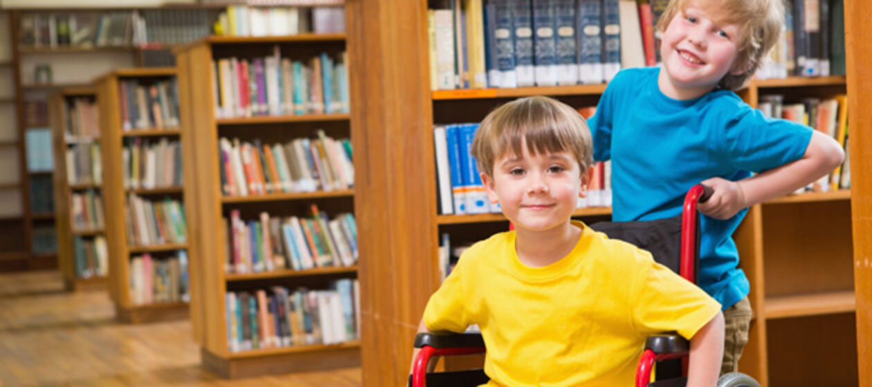 Ein Junge schiebt einen anderen im Rollstuhl durch Bücherei