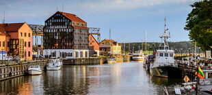 Der alte Hafen in Klaipeda (Litauen) mit kleinen Fischerbooten, Fachwerkhäusern, einem Baum, dem Hafenbecken und Wäldern der kurischen Nehrung im Hintergrund