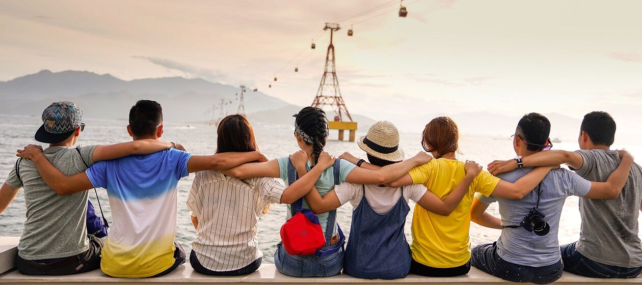 Eine Gruppe Jugendlicher von hinten fotografiert sitzt Arm in Arm und schaut auf einen See, über den eine Seilbahn gespannt ist 