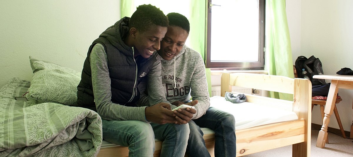 Zwei junge Geflüchtete sitzen in einem Zimmer auf dem Bett und schauen auf das Display eines Handys