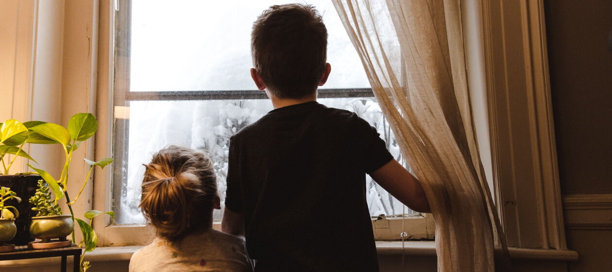 Zwei Kinder schauen aus dem Fenster einen verschneiten Baum an 