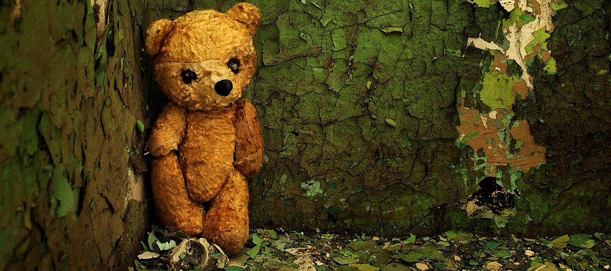 Ein alter Teddybär steht verlassen in der Ecke einer dreckigen, dunklen Zimmerwand.