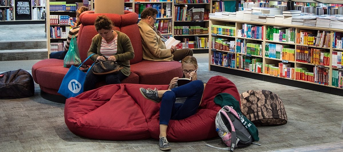 Personen lesen in einer Buchhandlung, im Vordergrund sitzt ein Mädchen auf einem Sitzsack