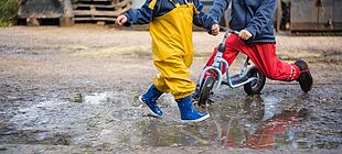 Zwei Kinder mit Regenhose und Gummistiefeln spielen in einer Pfütze