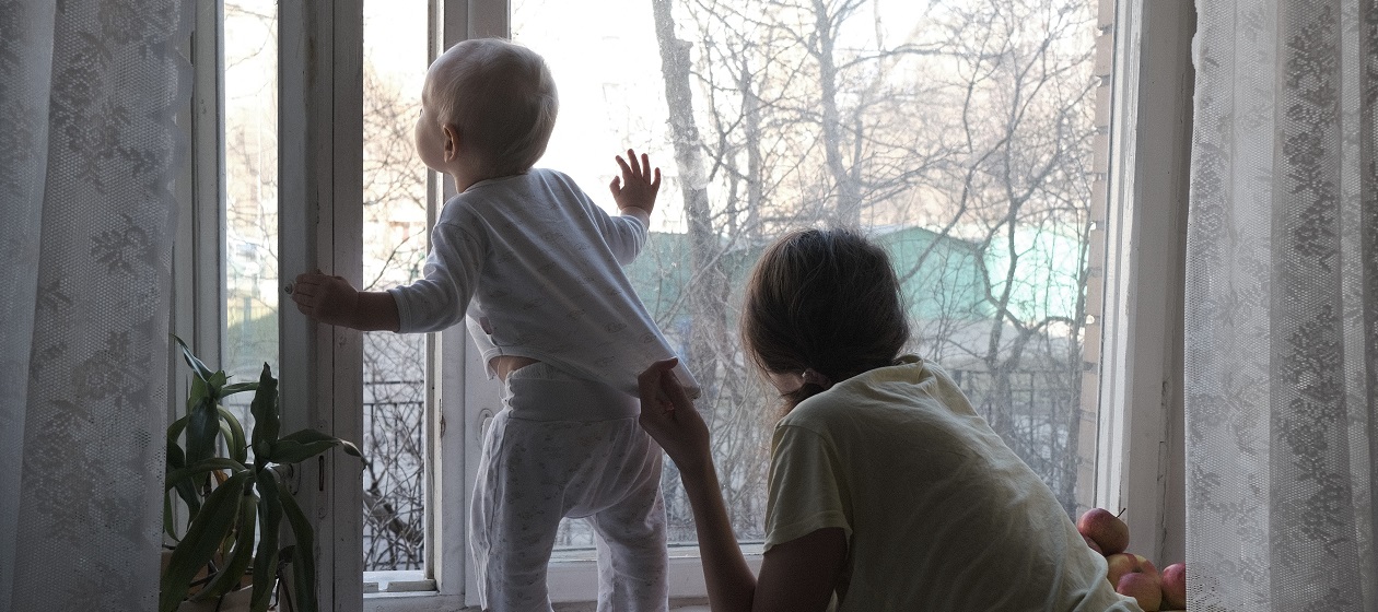 Ein Kleinkind steht an einem offenen Fenster und schaut nach draußen, eine erwachsene Person hält es von hinten fest