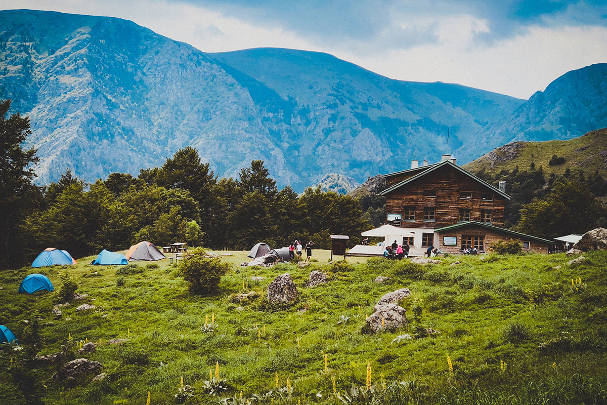 Jugendliche zelten auf einer Wiese vor einem Landhaus mit Alpenpanorama